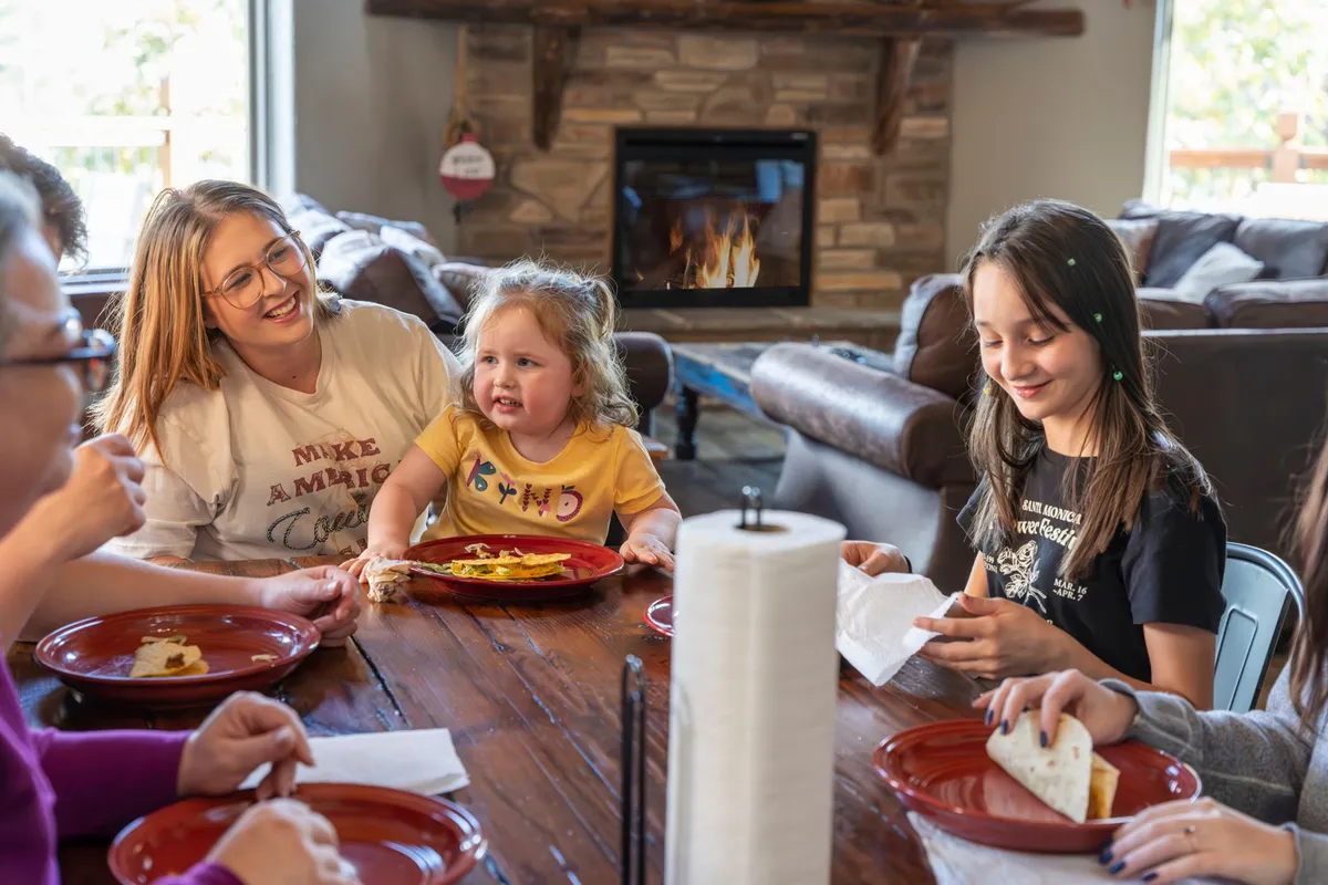 A joyful family scene with people gathered around a dining table sharing a meal, with a fireplace glowing in the background and food on the table. The mood is festive, relaxed, and full of laughter.