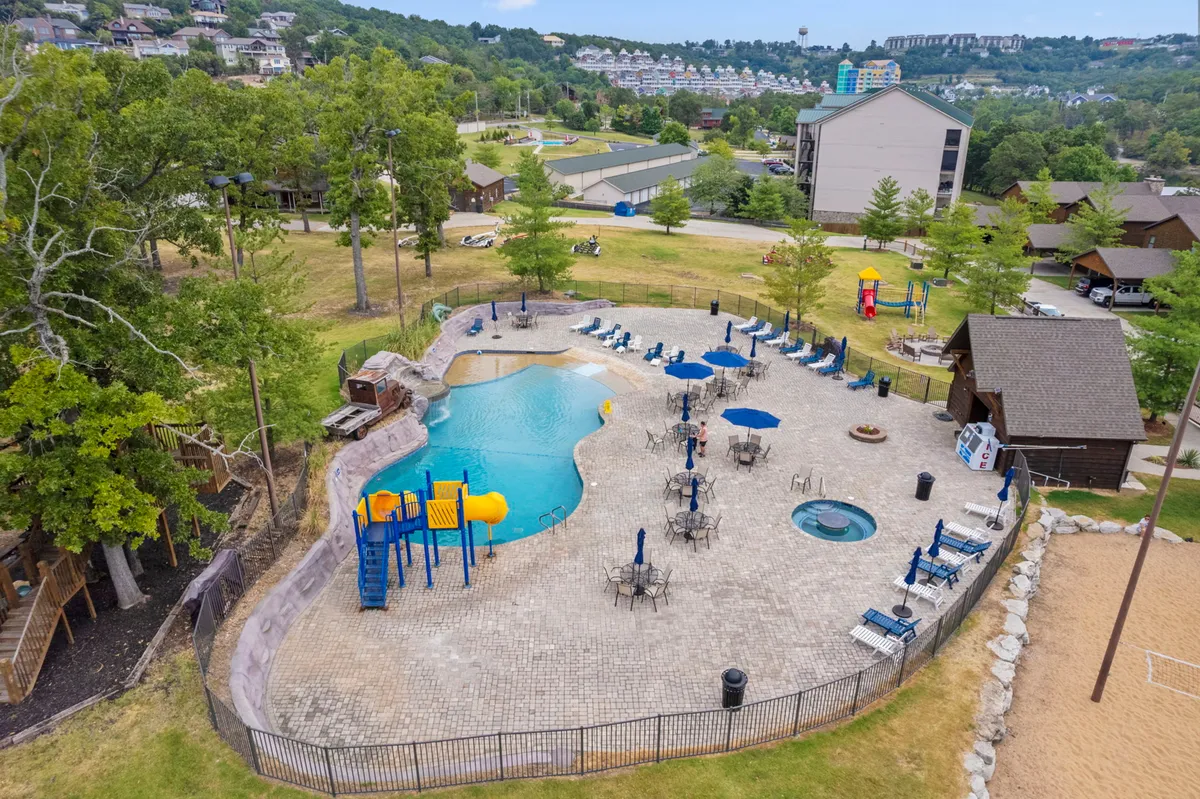 A vibrant scene shows the resort's beach-entry swimming pool and adjacent hot tub, surrounded by lounge chairs, a playground, and a sand volleyball court. Families and friends can be seen enjoying various activities, highlighting the lively, communal atmosphere.