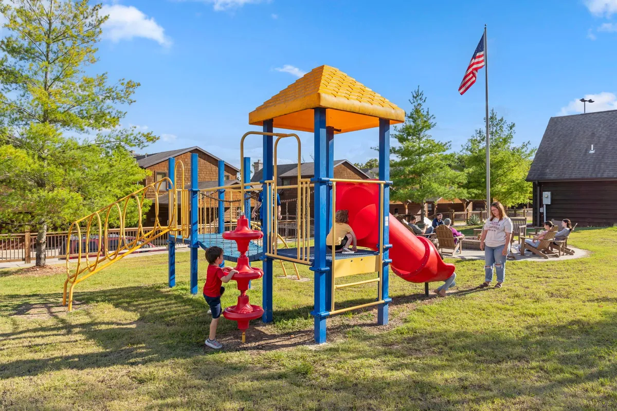 The photo captures children playing on a colorful playground structure while adults watch from nearby seating areas. The atmosphere is vibrant and communal, illustrating the safe, dedicated play spaces where kids can have fun and parents can socialize or relax.