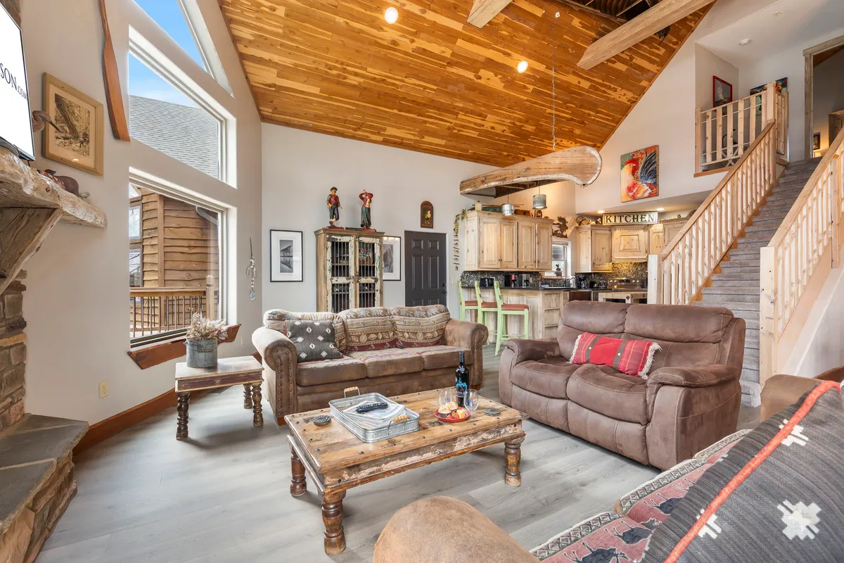 The image showcases a spacious, inviting living room with a soaring stone fireplace as the centerpiece, flanked by large windows that flood the space with natural light. Exposed wooden beams run overhead, and plush, comfortable sofas encircle a rustic coffee table, setting the scene for cozy family gatherings or relaxing evenings by the fire.