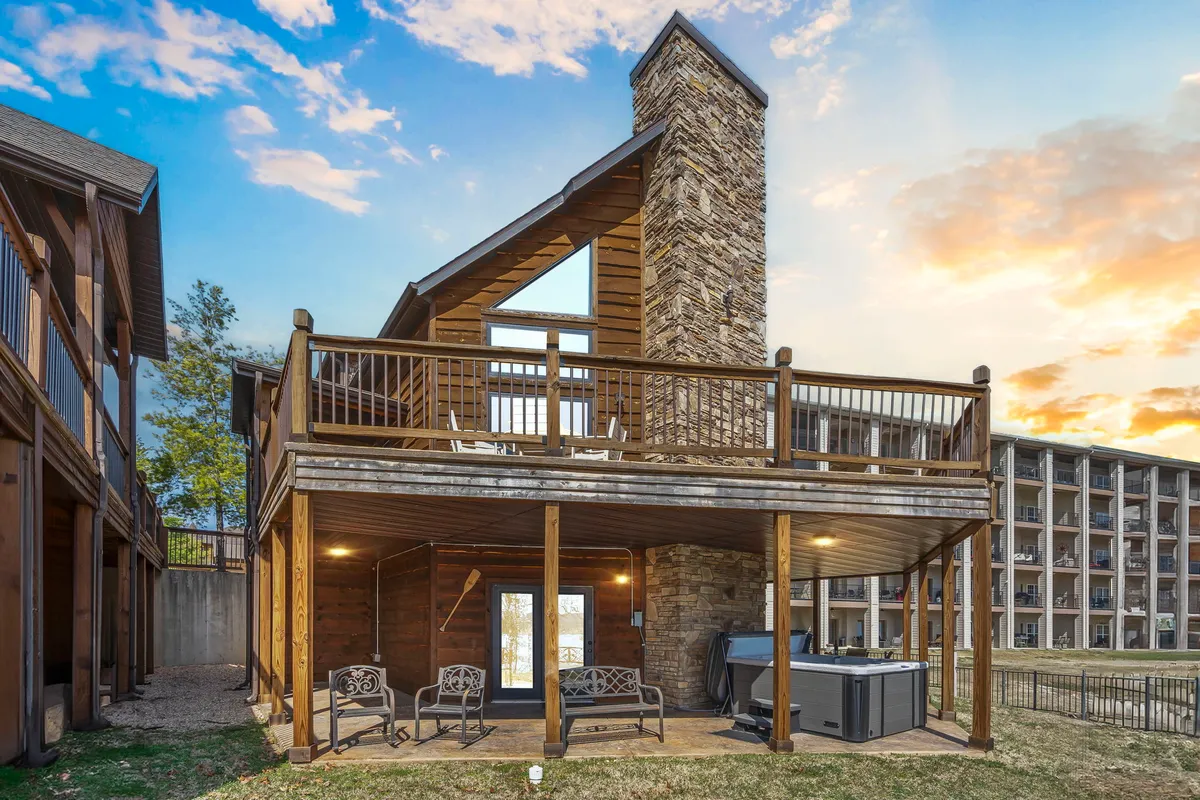 This exterior image captures a stone chimney flanking an expansive deck area, complete with a hot tub nestled under wooden railings. Large windows reflect the natural light, and there's space for outdoor gatherings, relaxing in the hot tub, or reading on the deck. The scene is set during sunset, enhancing the warm, inviting atmosphere.