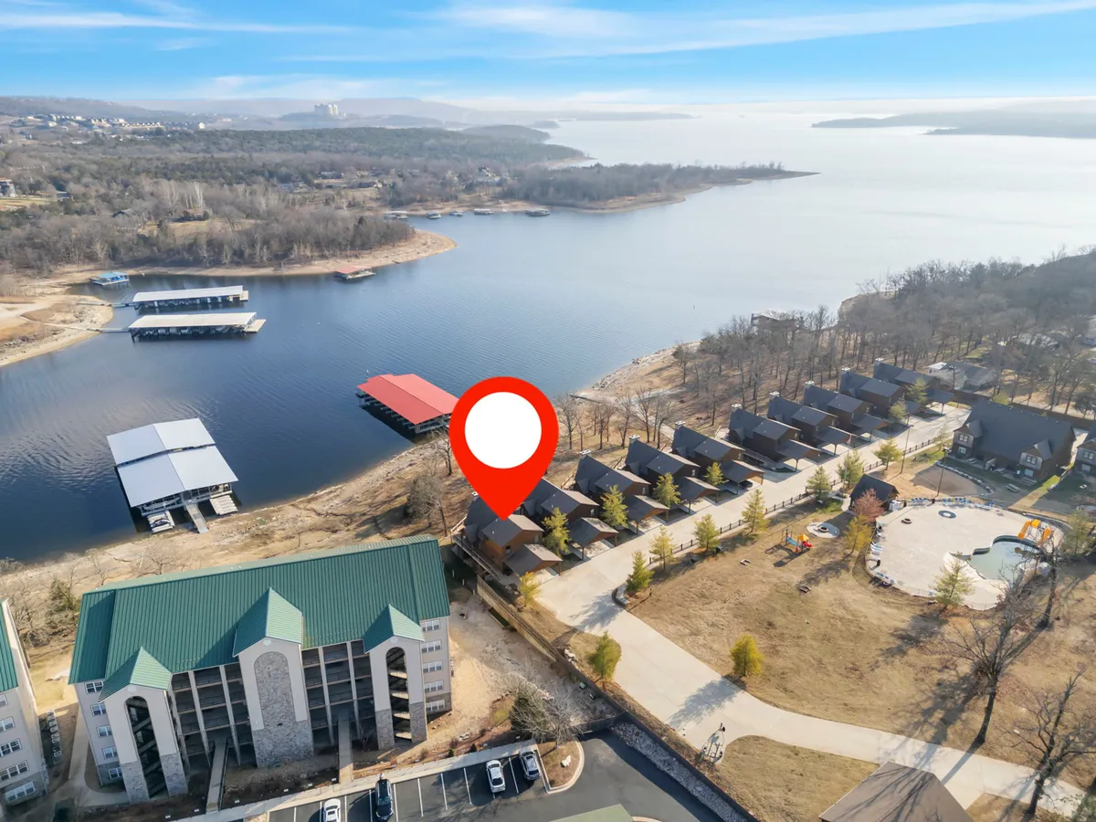 A serene aerial view showcases the lake, dock, cabins, playground, and distant mountains. This perspective highlights the integration of resort accommodations with the natural landscape, inviting guests and their pets to discover both relaxation and adventure along the shoreline.