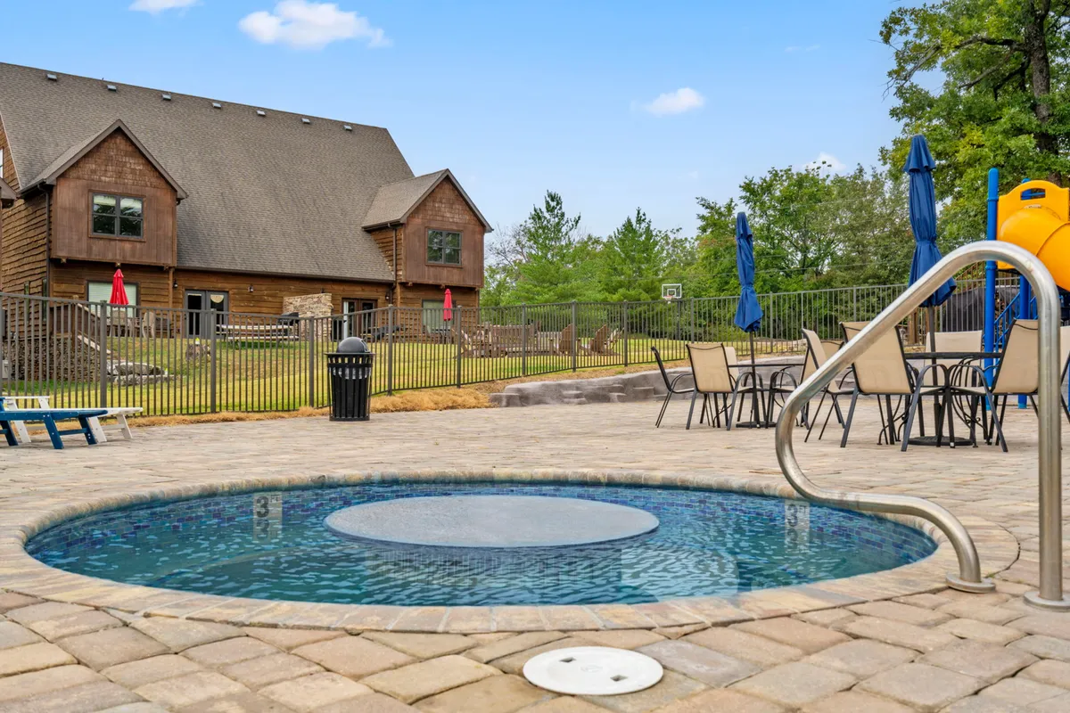 This image features a vibrant backyard with a swimming pool, a hot tub, patio seating, and a playground slide, all set against lush greenery. The inviting pool and hot tub are focal points, with the pool reflecting the open sky and the hot tub set beside the patio, suggesting relaxation and recreation for all ages. The presence of the slide and outdoor furniture hints at lively summer gatherings and family fun.