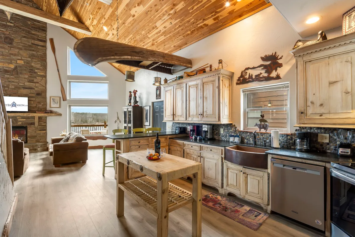 A welcoming open-concept space featuring a kitchen island with stools, a stone fireplace, large windows offering scenic views, and a distinctive wooden canoe hanging from the ceiling. The area is filled with natural light and seamlessly connects the kitchen to the living space.