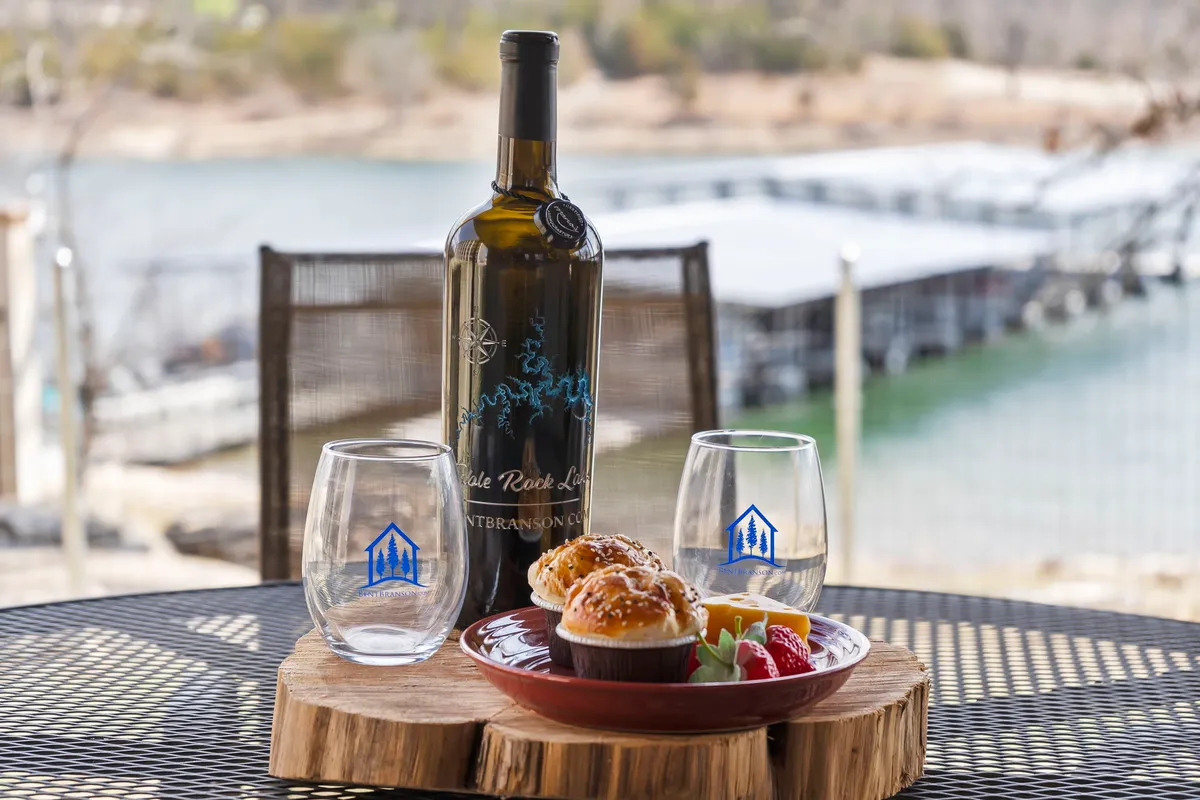 A cheerful lakeside setting with a bottle of wine, wine glasses, and a plate of muffins and fruit set on a table, all overlooking a sparkling water view. The scene conveys leisurely enjoyment and memorable moments shared with friends or loved ones.
