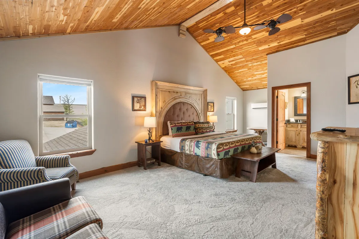 A spacious king-sized bed with a decorative headboard sits beneath rustic wooden ceiling beams. A striped armchair is positioned near a window that bathes the room in soft natural light, creating a restful, private retreat.