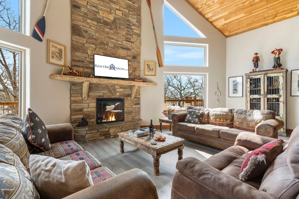 A cozy living area centered around a stone gas fireplace with comfortable sofas and large windows framing tranquil lake views. The wooden coffee table is perfect for board games or casual chats, and the setting radiates warmth and hospitality.