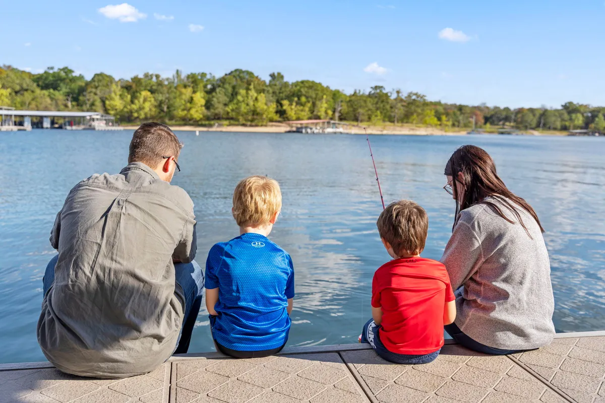A joyful family relaxes by the water's edge, with children fishing beside the calm lake. The scene captures the simple pleasures of lakeside living, from fishing to family bonding and enjoying nature.