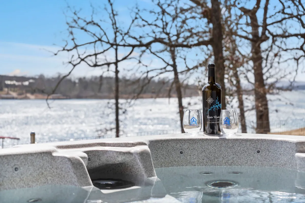 An outdoor hot tub sits on a patio overlooking the lake, with a wine bottle and glasses set nearby. The scene suggests relaxation and indulgence under the open sky, framed by natural beauty and trees.