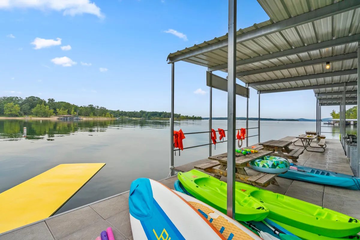 The photo shows colorful kayaks lined up on a dock overlooking a peaceful lake with picnic tables nearby. The calm water and inviting outdoor setup set the scene for kayaking, sunbathing, picnicking, and fishing. The image conveys a sense of readiness for adventure and relaxation.