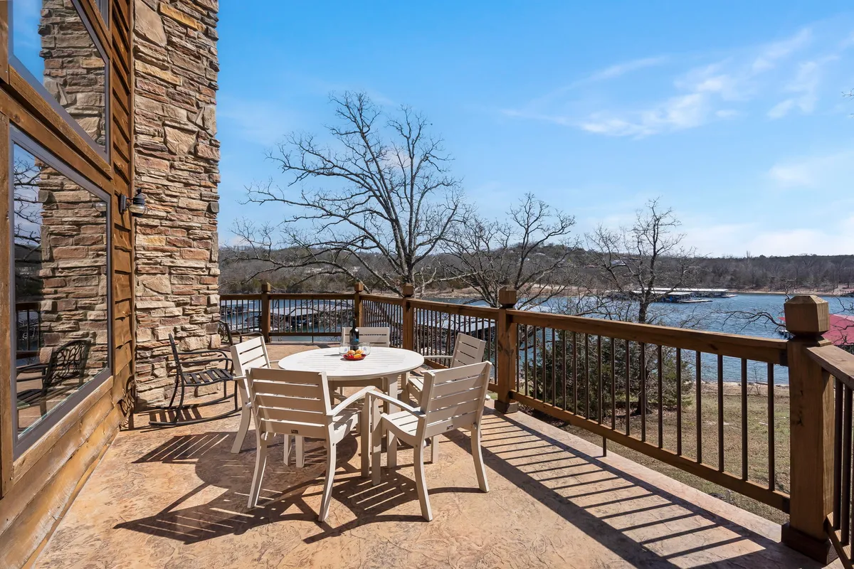 A serene outdoor dining table set on a wooden deck overlooks the tranquil lake, framed by a wooden railing. The scene invites guests to savor meals or relax with a drink while enjoying the peaceful water view and natural surroundings.